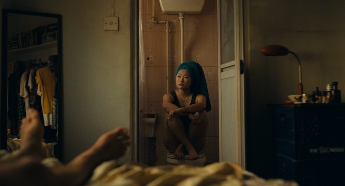 An Asian woman sits on a closed toilet with her legs up, smoking a cigarette in a pink bathroom with the door open and her hair wrapped up in a teal towel.