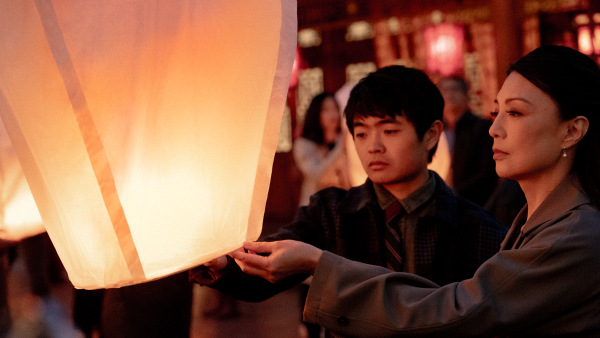 Li Fong (Ben Wang) and Dr. Fong (Ming-Na Wen) solemnly release a glowing sky lantern together during a nighttime ceremony. Their faces are softly lit by the warm orange light, surrounded by a crowd in the background amid red lanterns and traditional architecture.