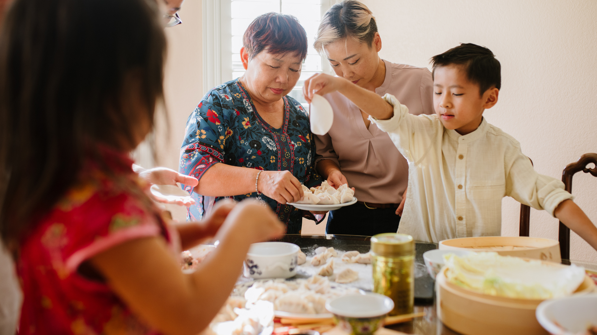 A multigenerational Asian American family gathers around a table making dumplings together, with adults and children participating in the food preparation, highlighting the warmth and connection of shared family traditions.