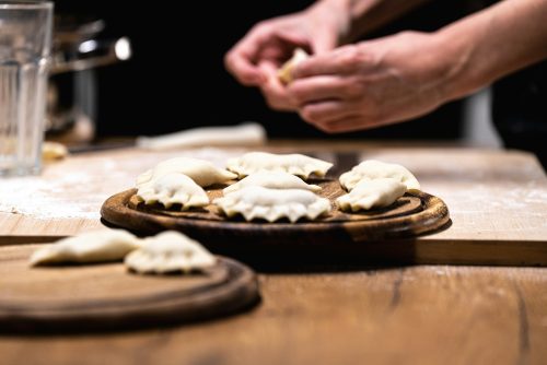 A plate of dumplings is in the foreground, while hands in the background are making more dumplings.