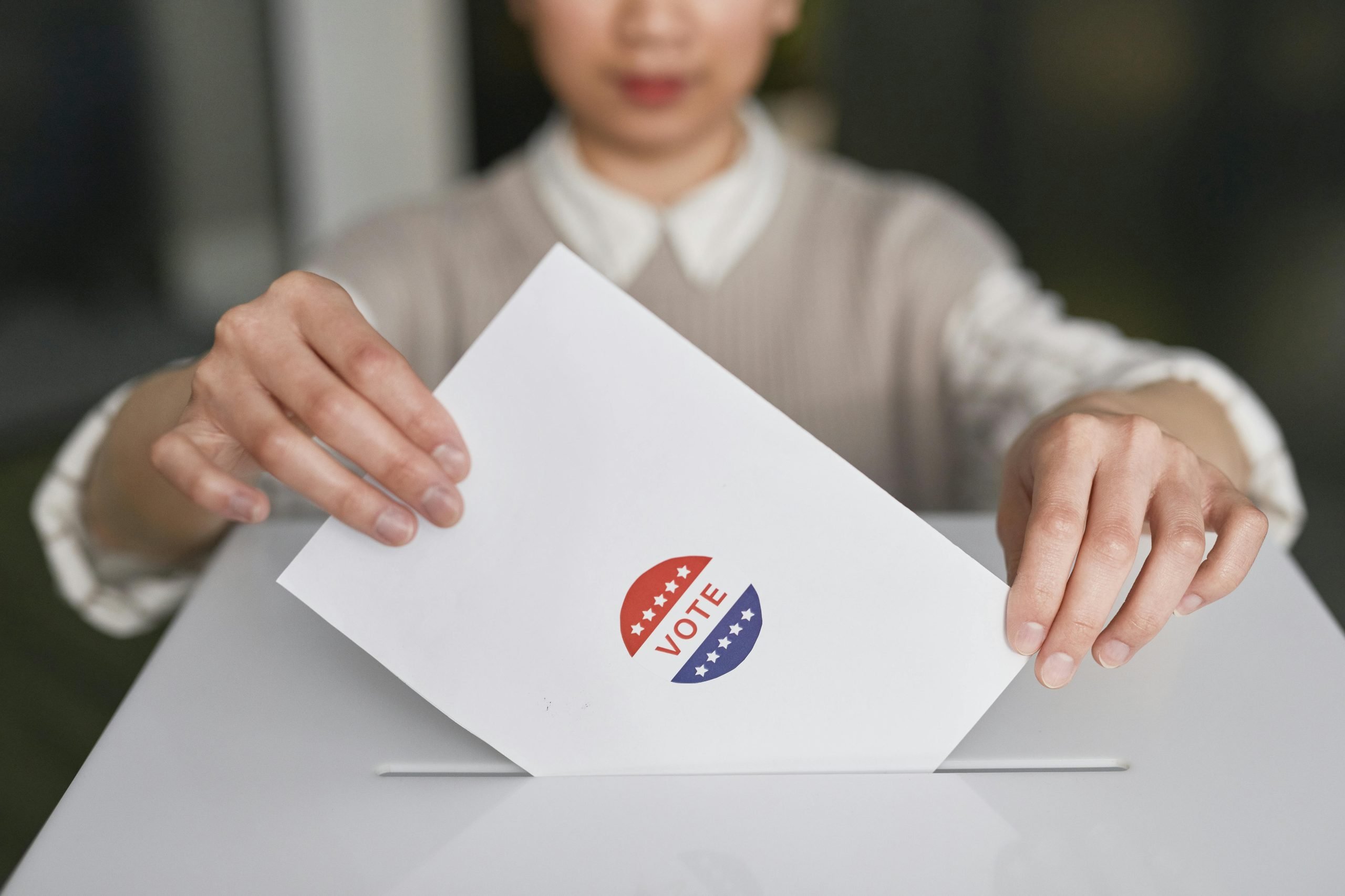 The lower half of an Asian woman's face is visible as she places a white paper with red, white, and blue sticker label reading "VOTE" into a white box.