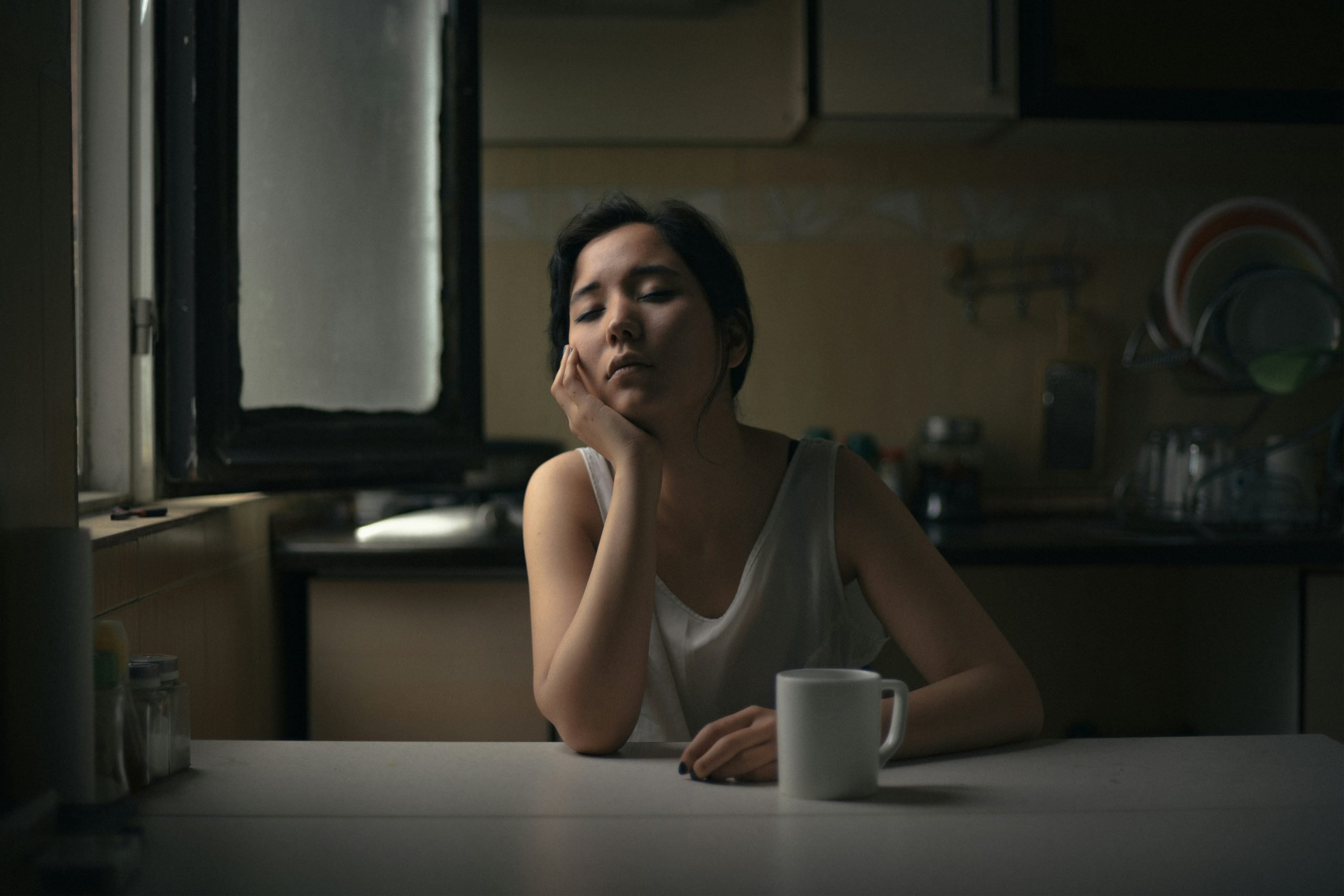 A middle-aged Asian woman sitting at a kitchen table, resting her chin on her hand beside a mug, appearing tired and contemplative in soft morning light.