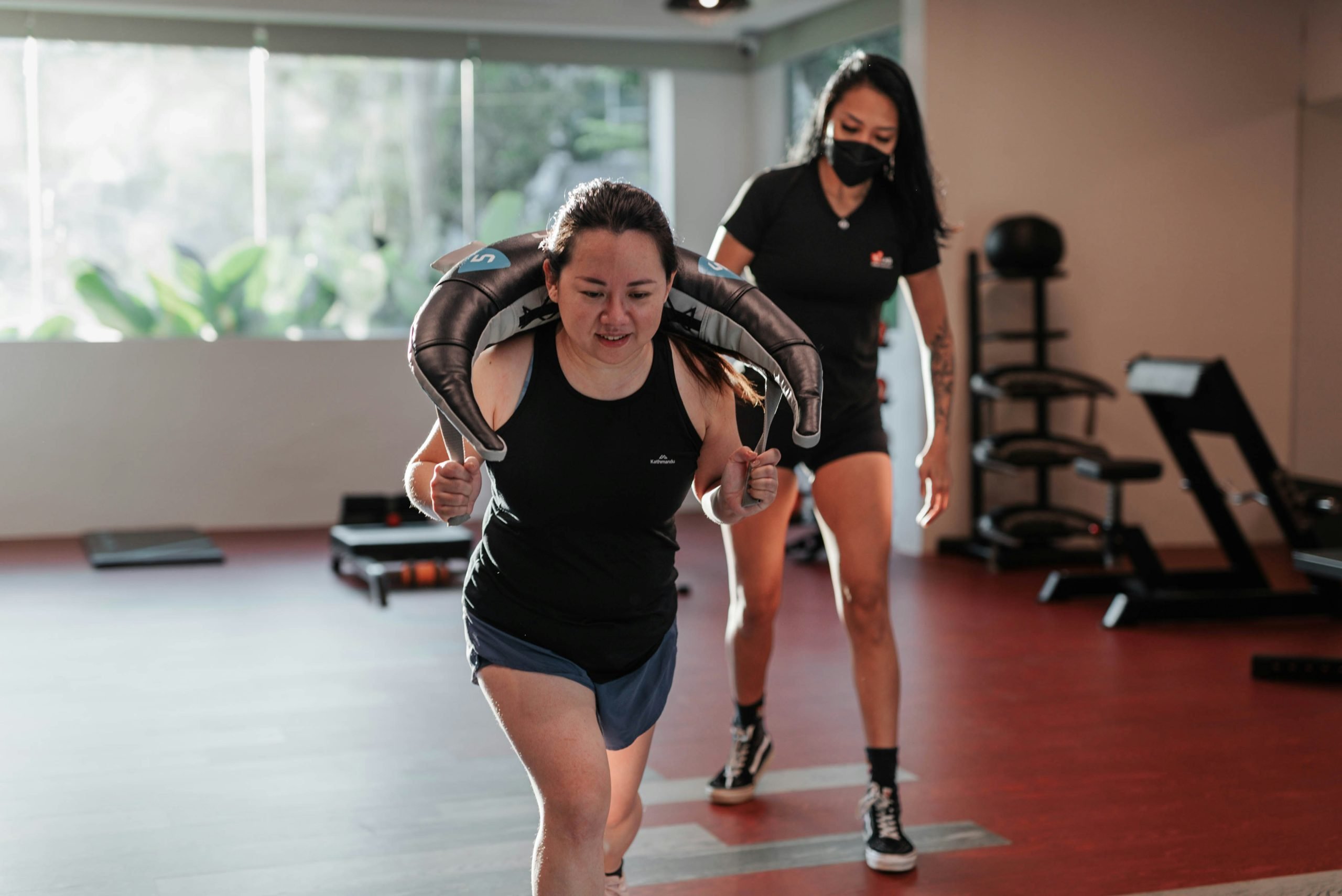 An Asian woman exercising in a gym with the guidance of a female trainer, carrying a weighted sandbag on her shoulders while walking forward.