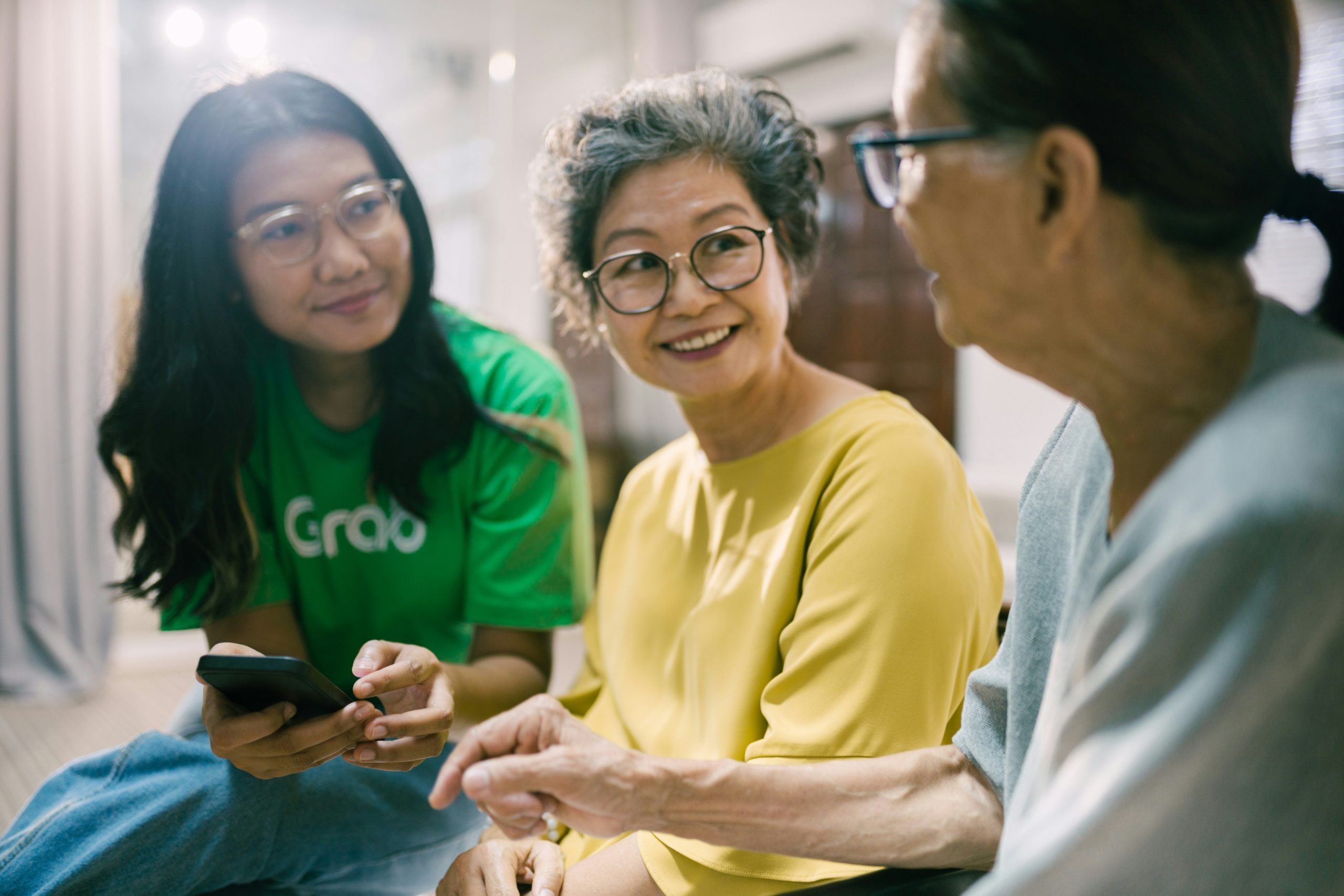 Three Asian women of different generations sitting together, smiling warmly as they talk, one holding a smartphone while the others listen.