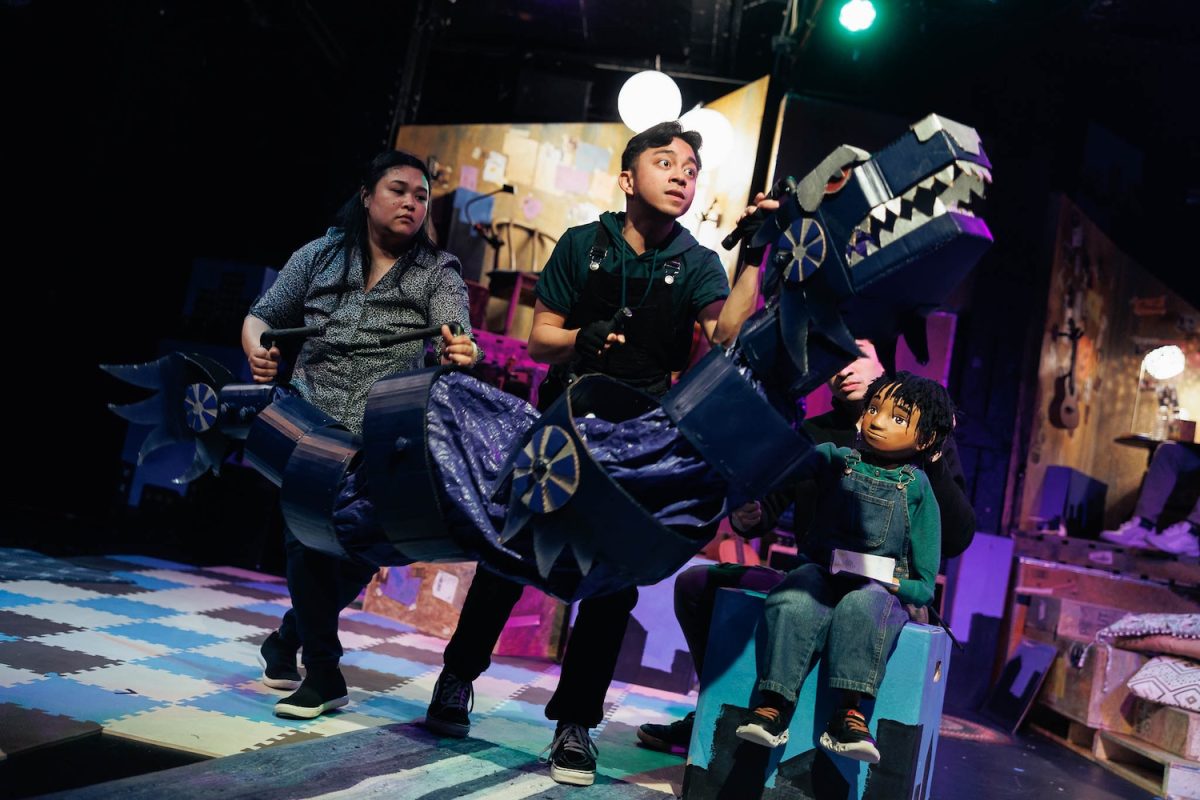 On a theater stage, two Asian American performers operate a large, segmented blue dragon puppet with silver accents. To the right, another performer sits next to a young boy puppet wearing denim overalls and a green shirt. The set is eclectic, featuring wooden crates, a checkered floor, and warm, spherical stage lighting, capturing a whimsical scene from a CHUANG Stage production.