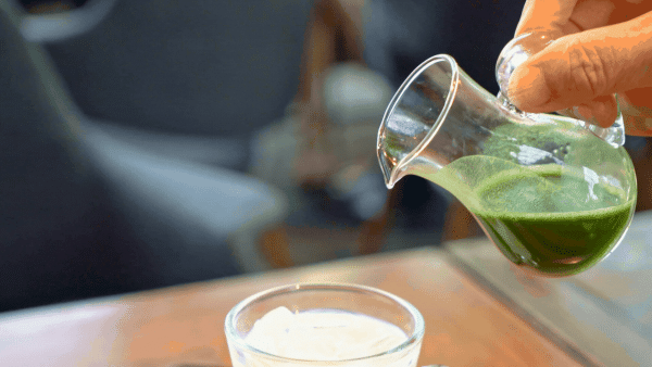 A close-up, slightly angled shot of a hand holding a small glass pitcher, pouring thick, vibrant green matcha into a clear glass filled with white milk and ice. The matcha swirls into the milk on a wooden table, creating a marbled effect.