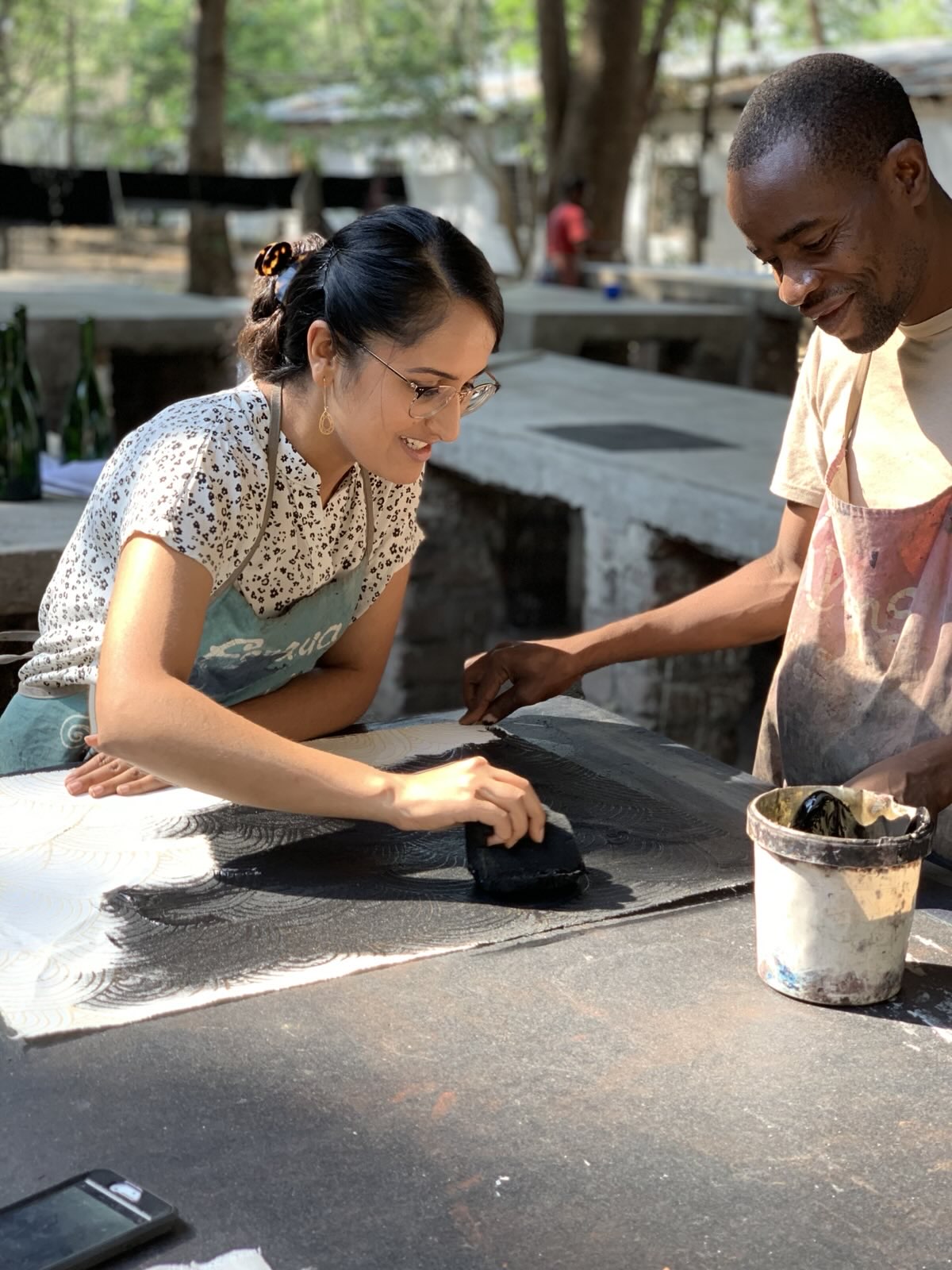 Manpreet Kaur Kalra wearing an apron and glasses, smiling as she collaborates with an artisan on a black and white textile print in an outdoor workshop.