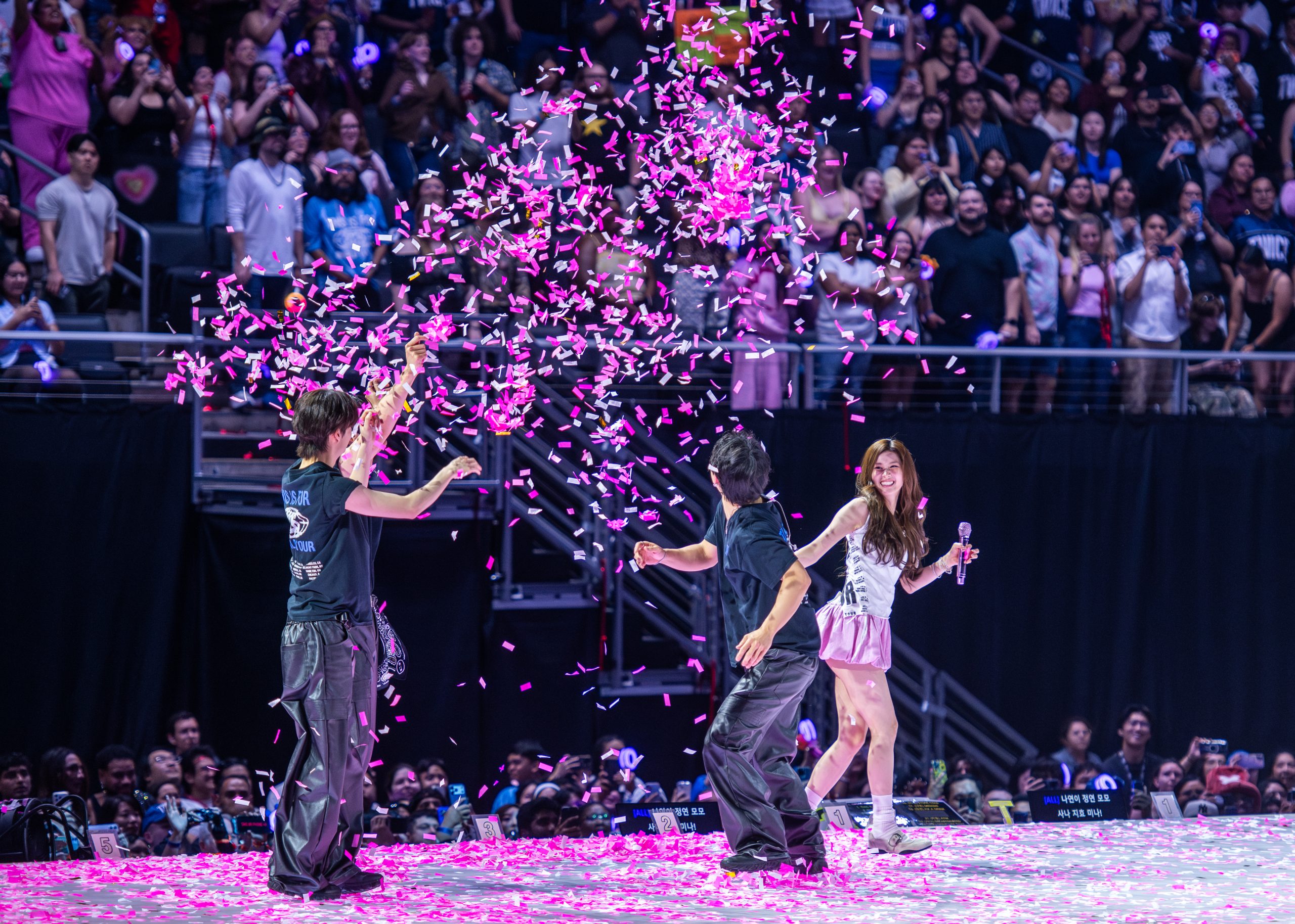 Two members, one in a long high ponytail and the other with her dark black hair down, walk across the stage, smiling and waving to the audience with their arms raised high. The two wear matching blue, black and white stage costumes.