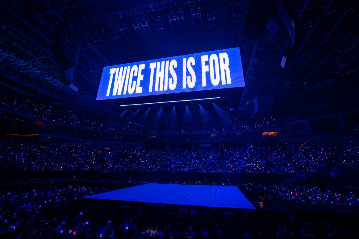 A wide shot of a darkened arena filled with a crowd of glowing blue lightsticks. A massive hanging screen displays the words "TWICE THIS IS FOR" in bold white letters.
