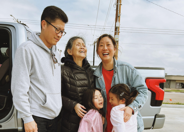 A group of five Asians stand close together on a driveway: a man with short black hair, glasses, gray sweatshirt, and black pants stands on the far left next to a laughing elderly woman in a black puffy jacket and black pants. To their right is a mid-life woman with brown hair pulled back and a jean jacket over a red shirt. She is hugging two young girls close to her, both with brown hair, pink long-sleeved shirts, and brown and peach pants.