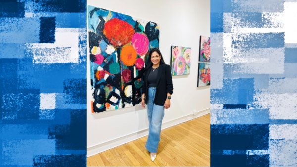 A woman stands smiling in an art gallery beside a large, colorful abstract floral painting. She wears a black blazer, dark top, wide-leg jeans, and white pointed shoes, with several vibrant paintings displayed on the white gallery walls behind her.