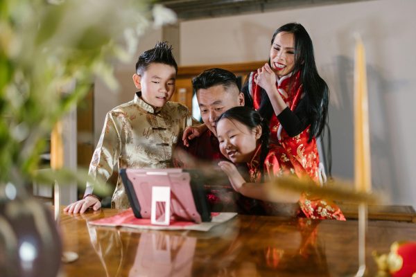 A Chinese family of four, consisting of a mom, dad, daughter, and son, are dressed in traditional gold and red silk outfits. They are huddled around a pink and black tablet that sits on a brown table.