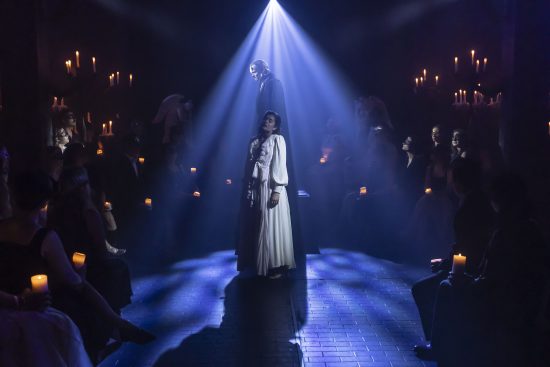 A woman in a flowing white dress stands beneath a bright blue spotlight at the center of a narrow aisle, while a cloaked figure looms behind them. Audience members seated on either side hold glowing candles and watch intently.