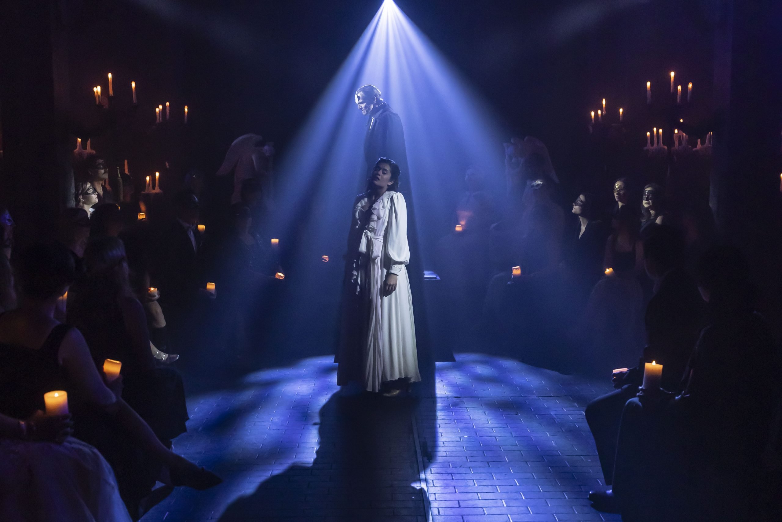 A woman in a flowing white dress stands beneath a bright blue spotlight at the center of a narrow aisle, while a cloaked figure looms behind them. Audience members seated on either side hold glowing candles and watch intently.