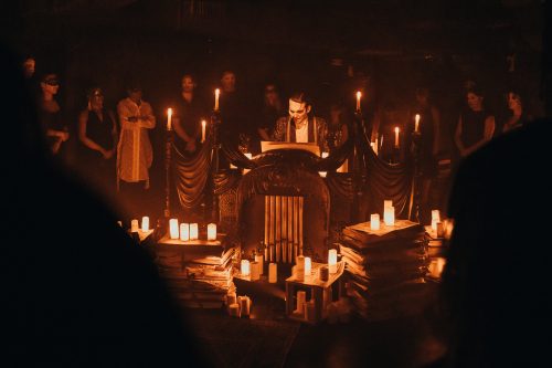 In a dimly lit room bathed in warm amber light, a masked man stands behind an ornate organ or lectern surrounded by stacks of papers and dozens of flickering candles. Guests dressed in dark attire and masquerade masks form a circle around the scene, watching as the performer appears to read or deliver a dramatic monologue.