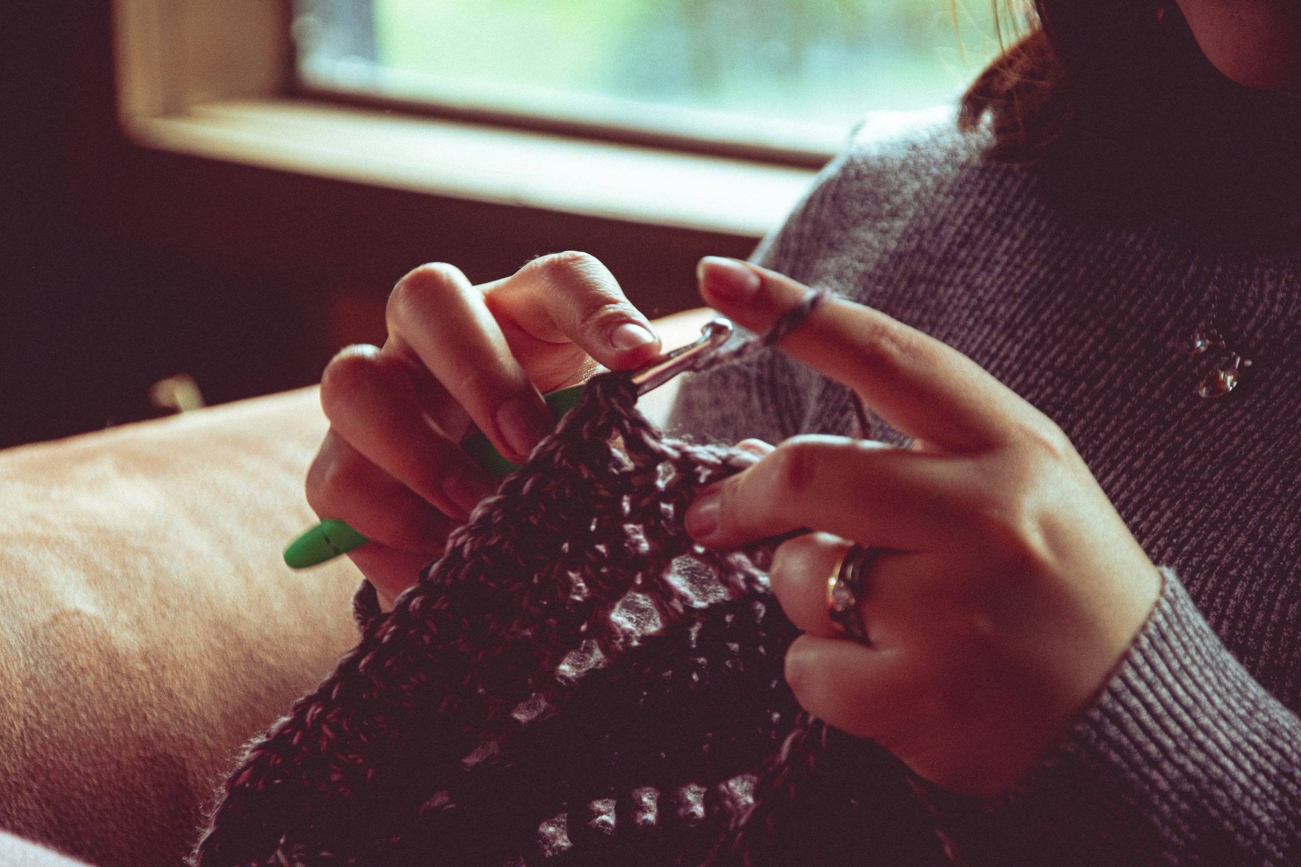 Close-up of a person crocheting a dark-colored project while seated indoors near a window. Their hands hold a crochet hook and yarn, with soft natural light illuminating the scene and creating a warm, cozy atmosphere. The person wears a gray knit sweater and rings on several fingers.