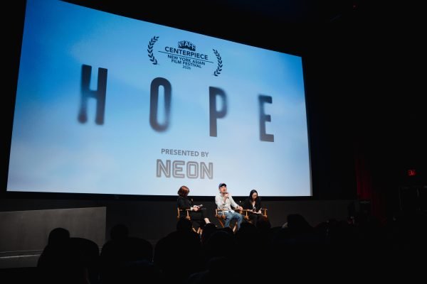 A man with dark-rimmed glasses, a blue baseball cap, and a light button-up shirt sits on a director's chair alongside two female panelists on a theater stage during a Q&A session. Behind them, a large screen displays the movie title "HOPE" beneath the New York Asian Film Festival logo.