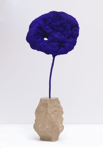 A sculpture consisting of a textured, cloud-like natural sponge dyed in intense ultramarine blue, mounted on a thin stem rooted in a rough stone pedestal.