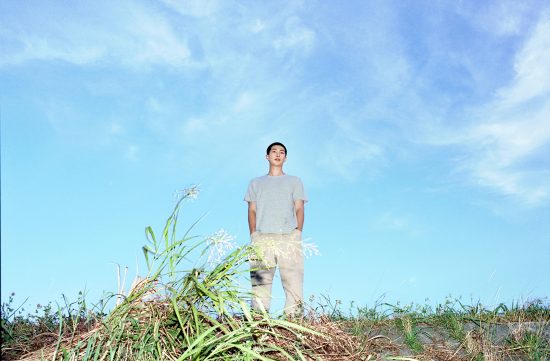A low-angle outdoor portrait of a young man with short dark hair wearing a plain grey t-shirt and beige pants, standing calmly against a vast blue sky behind tall wild grasses.