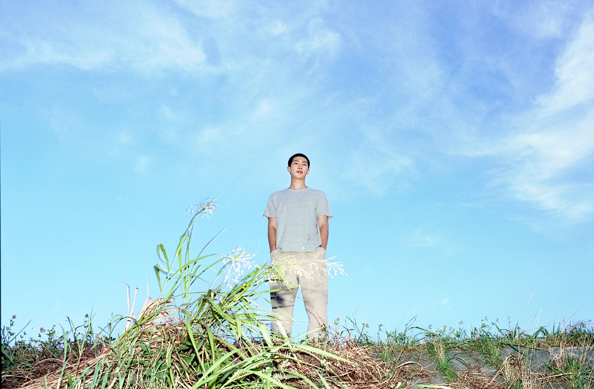 A low-angle outdoor portrait of a young man with short dark hair wearing a plain grey t-shirt and beige pants, standing calmly against a vast blue sky behind tall wild grasses.